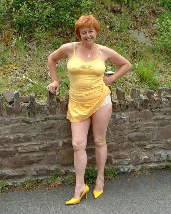 Femme rousse, portant une robe jaune, posant dehors, mains sur les hanches, souriante.