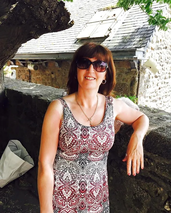 Femme aux cheveux bruns, en robe imprimée, souriant, portant des lunettes de soleil, appuyée contre un mur de pierre.