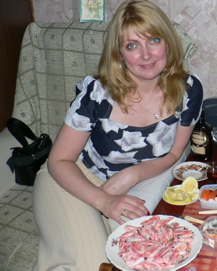 Femme aux cheveux blonds, en robe blanche, assise à table, souriant, devant une assiette de crevettes.