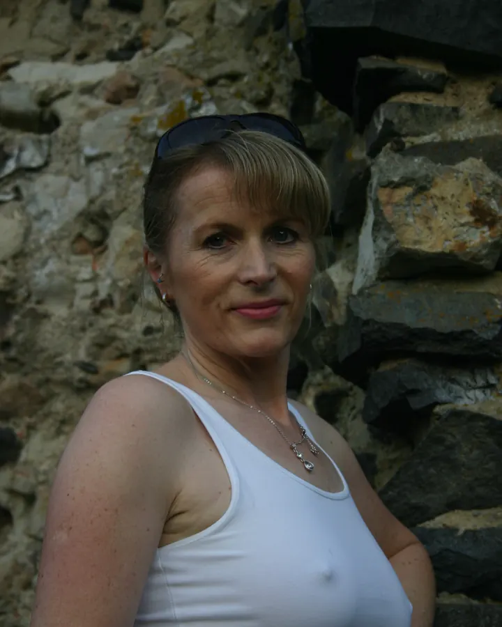 Femme aux cheveux bruns, en haut blanc, souriant, portant des lunettes de soleil, appuyée contre un mur de pierre.