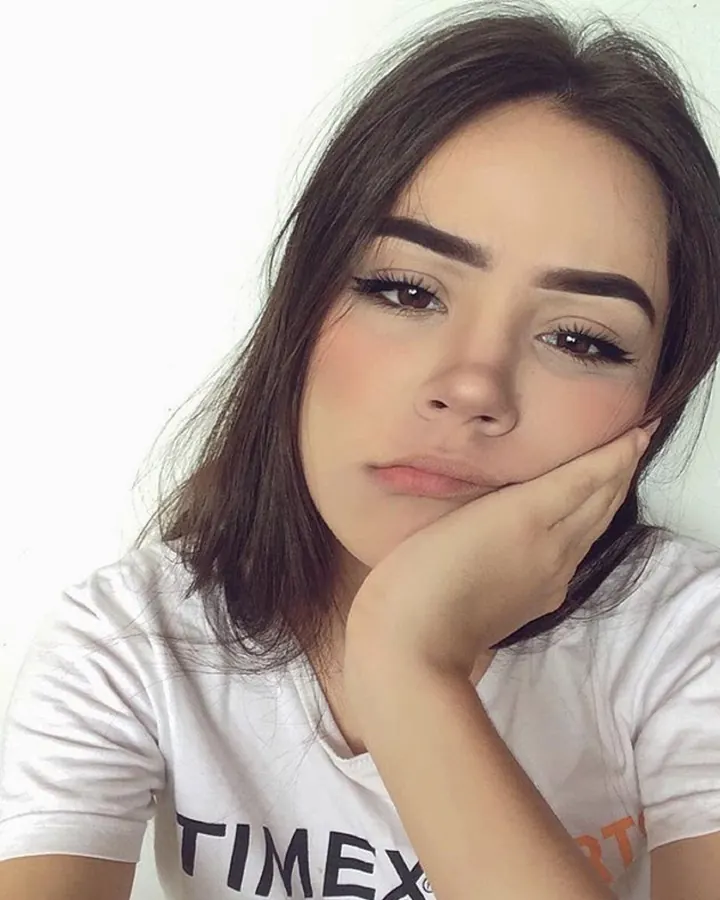 Jeune femme aux cheveux bruns foncés, vêtue d'un t-shirt blanc, posant le menton sur sa main, regard pensif.