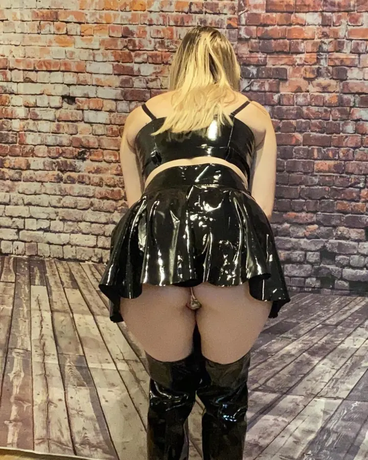 Femme blonde dos tourné, jupe et top latex noir brillant, bottes hautes