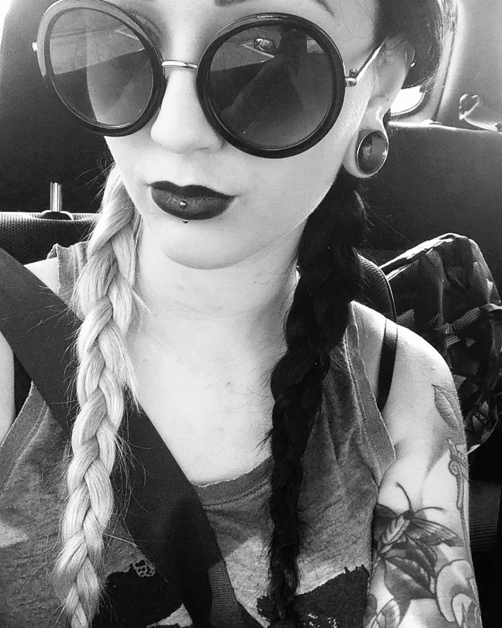 Jeune femme aux deux tresses, lunettes rondes, maquillage sombre, regard caméra
