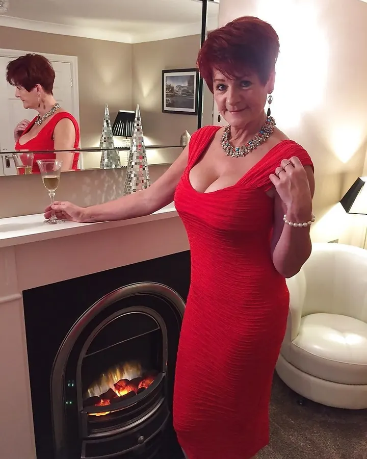Femme mature rousse en robe rouge, pose élégante près d’une cheminée