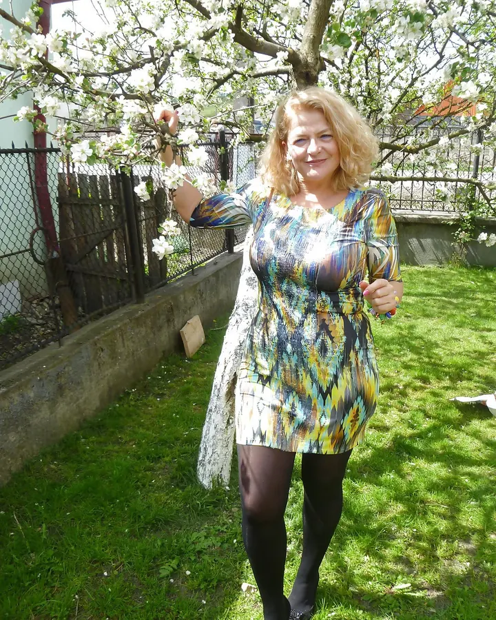 Femme blonde en robe colorée sous un arbre en fleurs, souriant à la caméra.