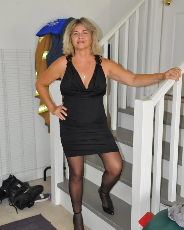 Femme blonde en robe noire, posant sur un escalier blanc, une main sur la hanche.