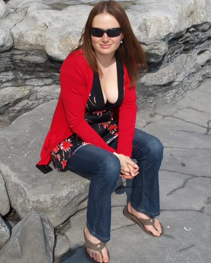 Femme brune en cardigan rouge et robe à fleurs, assise sur des rochers, souriant à la caméra.