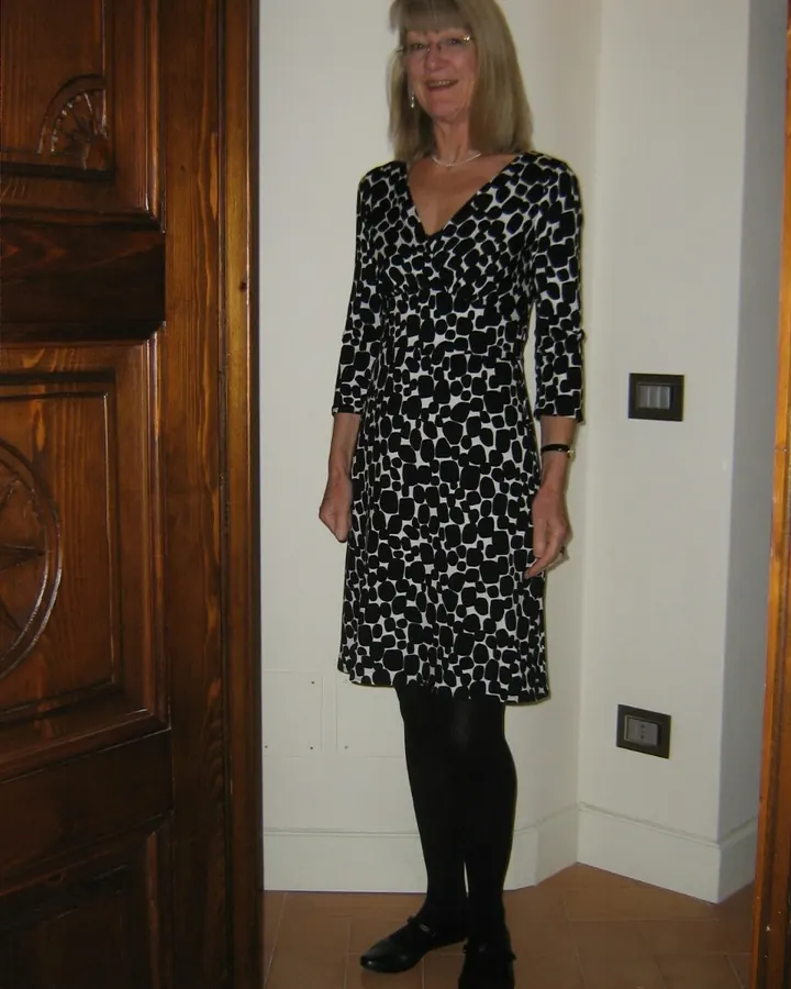 Femme aux cheveux blancs en robe à motifs noirs et blancs, debout près d'une porte en bois.