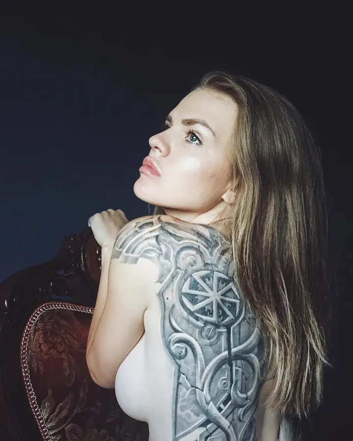 Femme blonde nue, montrant un tatouage complexe sur son épaule et son dos, assise sur une chaise ancienne.