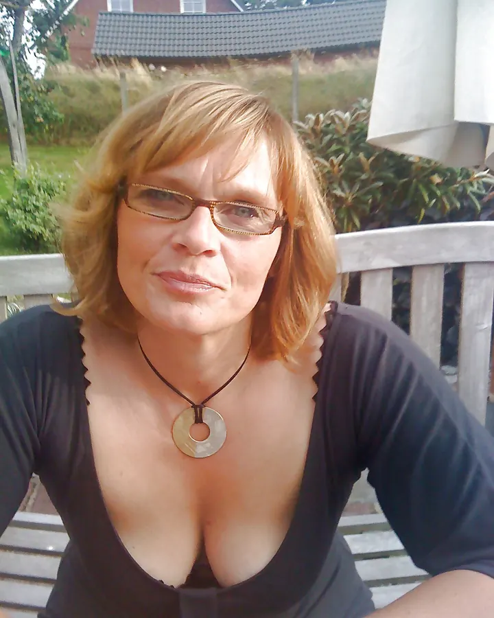 Femme aux cheveux blonds portant une robe noire, assise sur un banc en pierre extérieur, avec des lunettes de soleil.