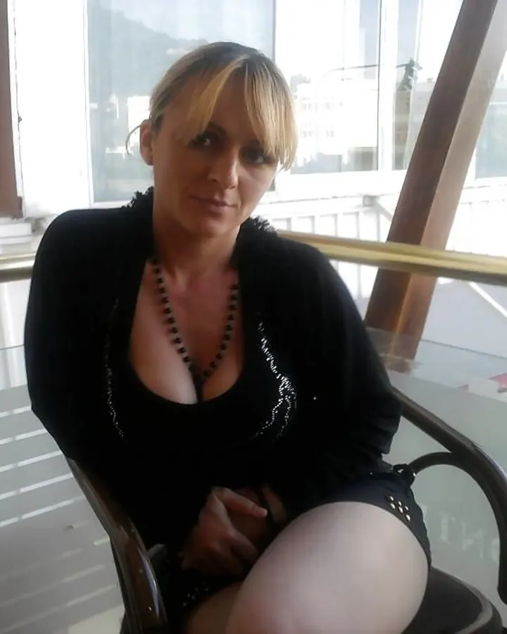 Femme aux cheveux blonds portant une robe noire, assise sur un banc en pierre extérieur, avec des lunettes de soleil.