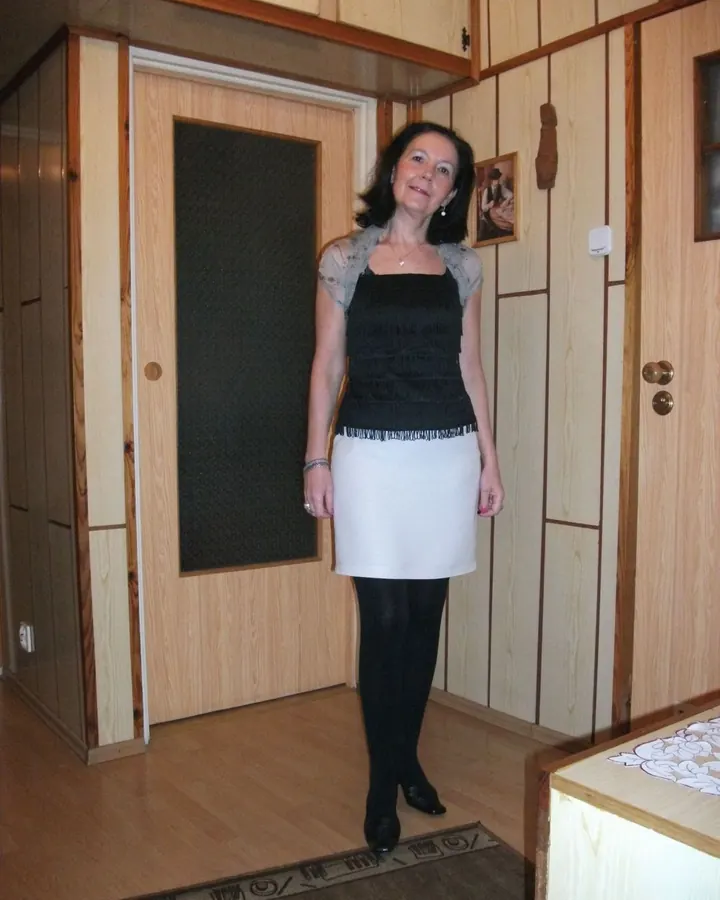 Femme aux cheveux noirs, assise dans une pièce en bois, portant une robe noire et blanche, regardant l'objectif avec un sourire discret.