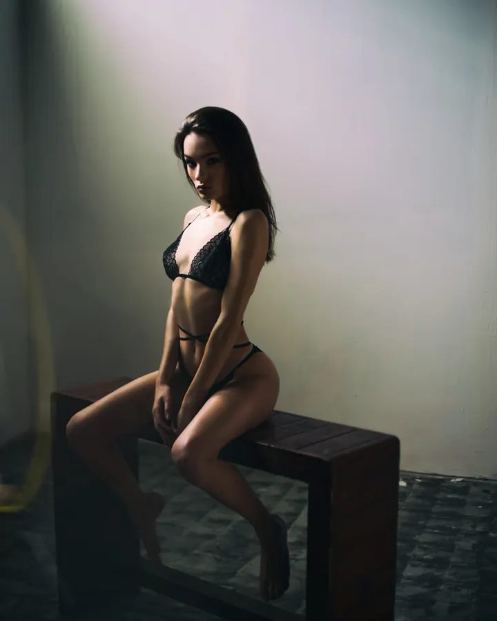 Femme indienne aux cheveux longs noirs, portant un ensemble de lingerie noire, assise sur un banc en bois, regardant l'objectif avec un sourire discret.