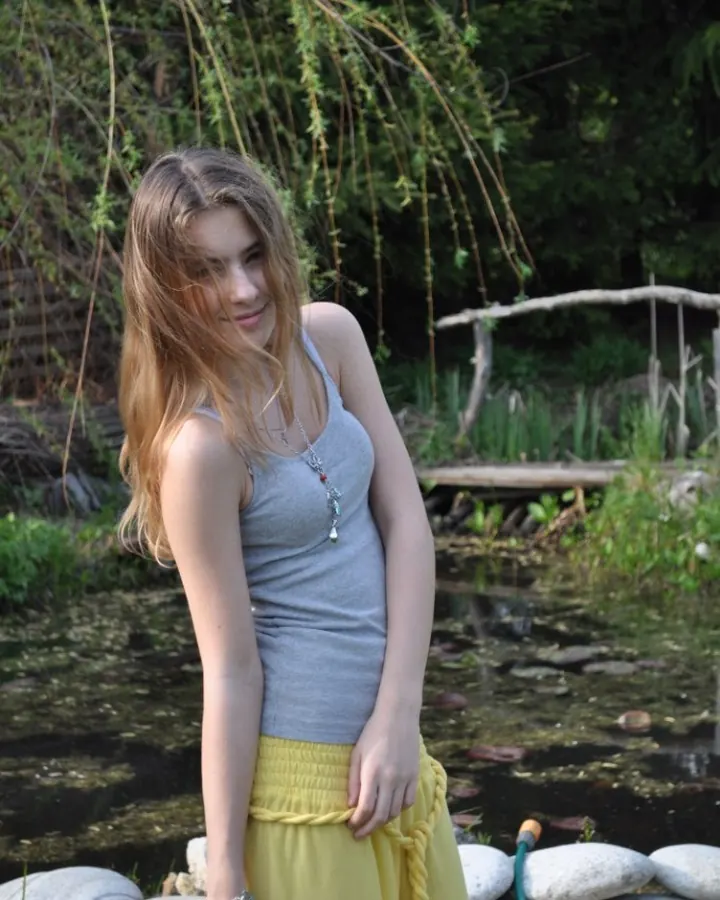 Jeune femme aux cheveux blonds, portant un haut gris et une jupe jaune, debout près d’un étang, regardant l’objectif avec un sourire discret.