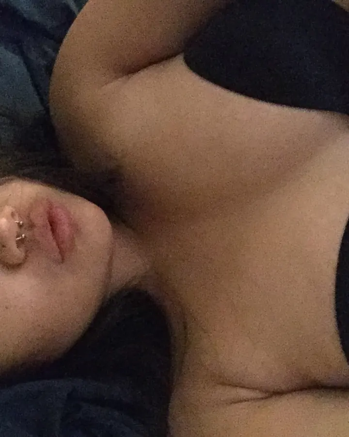 Femme aux cheveux foncés, allongée sur un lit sombre, portant un haut noir, regardant l'objectif avec un piercing au nez.