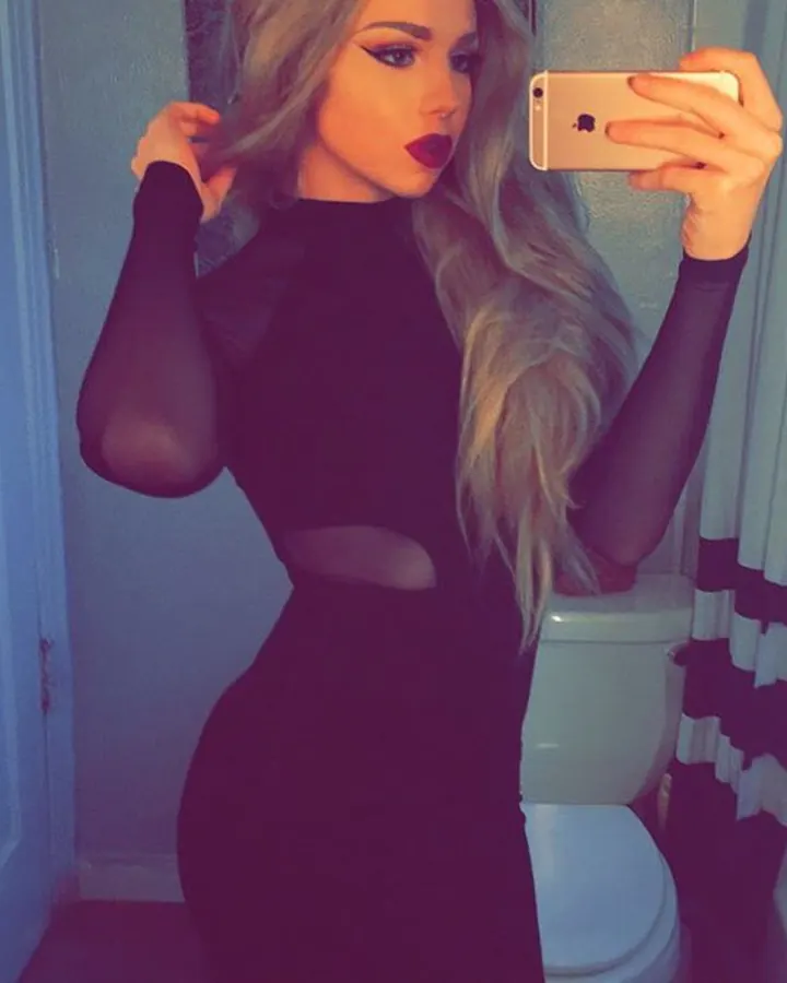 Femme en haut noir, prenant un selfie dans une chambre avec un lit et des couvertures colorées.