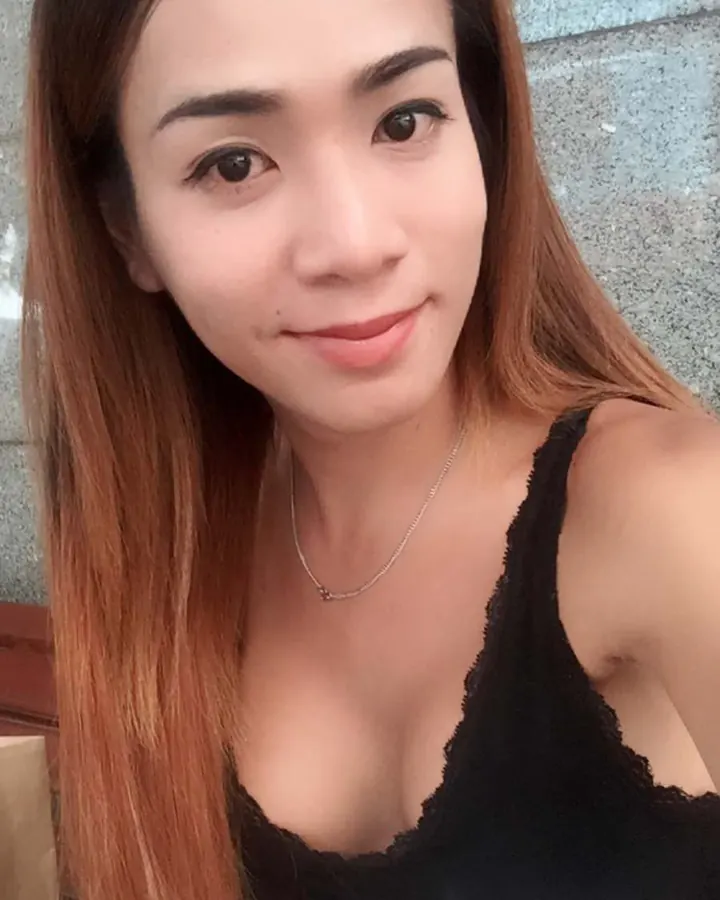 Femme souriante en haut noir, prenant un selfie en extérieur contre un mur gris.