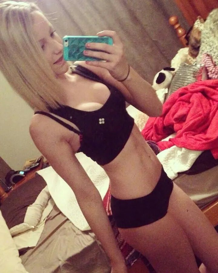 Femme en haut noir, prenant un selfie dans une chambre avec un lit et des couvertures colorées.