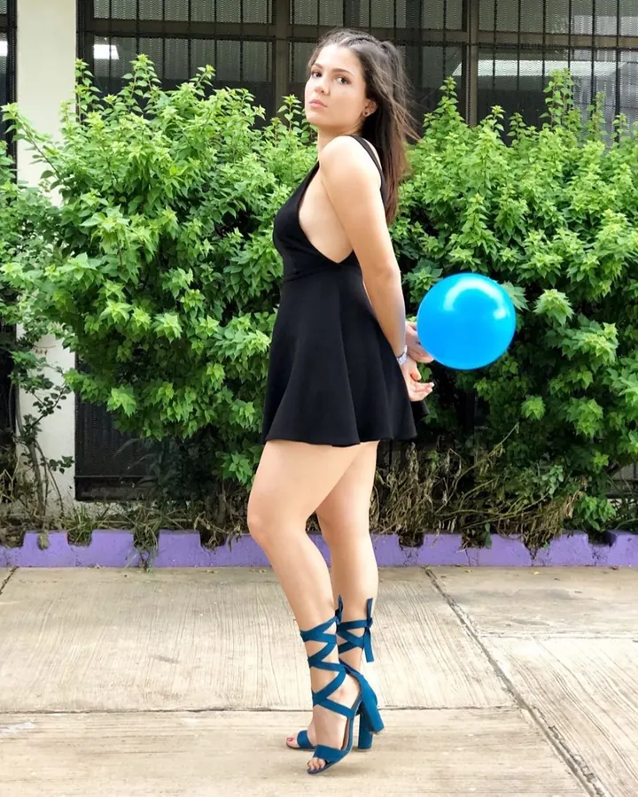 Femme en robe noire avec découpes au dos, tenant un ballon bleu, debout devant un feuillage vert.