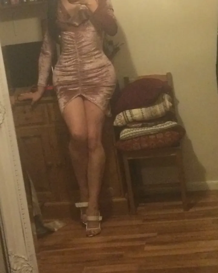Femme en robe rose velours, debout devant un miroir, avec une chaise et des coussins en arrière-plan.
