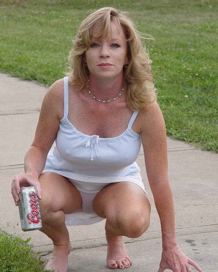 Femme blonde en tenue blanche accroupie sur un trottoir, tenant une canette de bière
