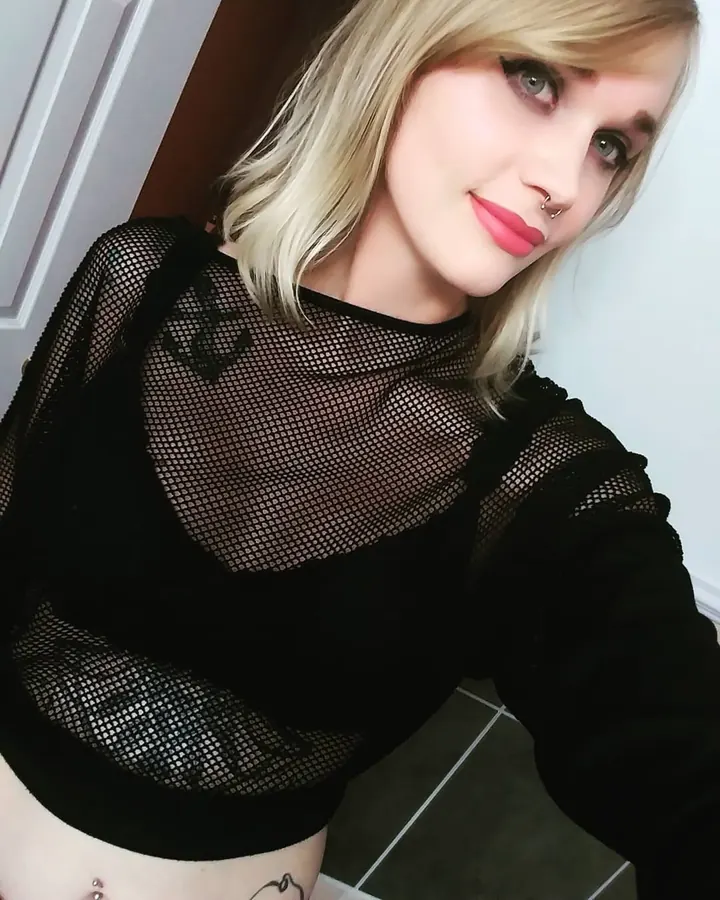 Femme aux cheveux blonds, nue du torse, portant un soutien-gorge noir, prenant une photo de profil devant un miroir.