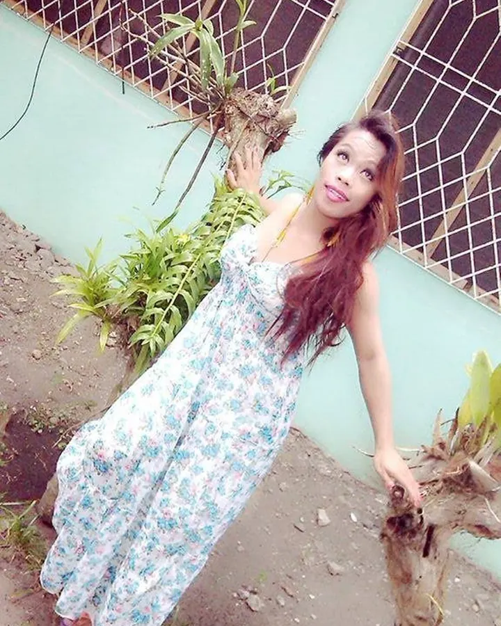 Femme aux cheveux bruns, portant une robe fleurie, posant dehors près d’un mur vert clair avec des plantes.