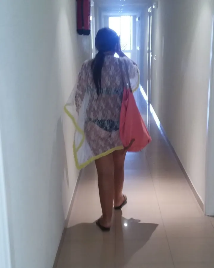 Femme vue de dos dans un couloir, portant une robe transparente à motifs et un sac rose, cheveux noirs en queue-de-cheval.