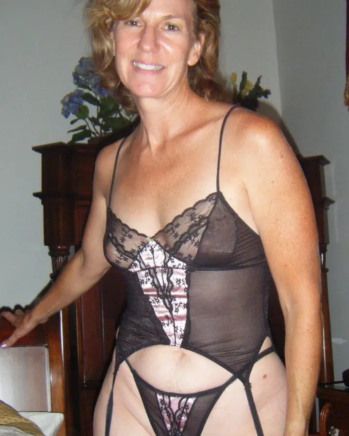 Femme aux cheveux blonds portant une robe noire avec dentelle, souriant à la caméra.