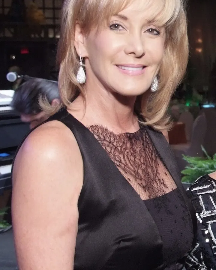 Femme aux cheveux blonds platine portant une robe noire avec dentelle, souriant à la caméra.