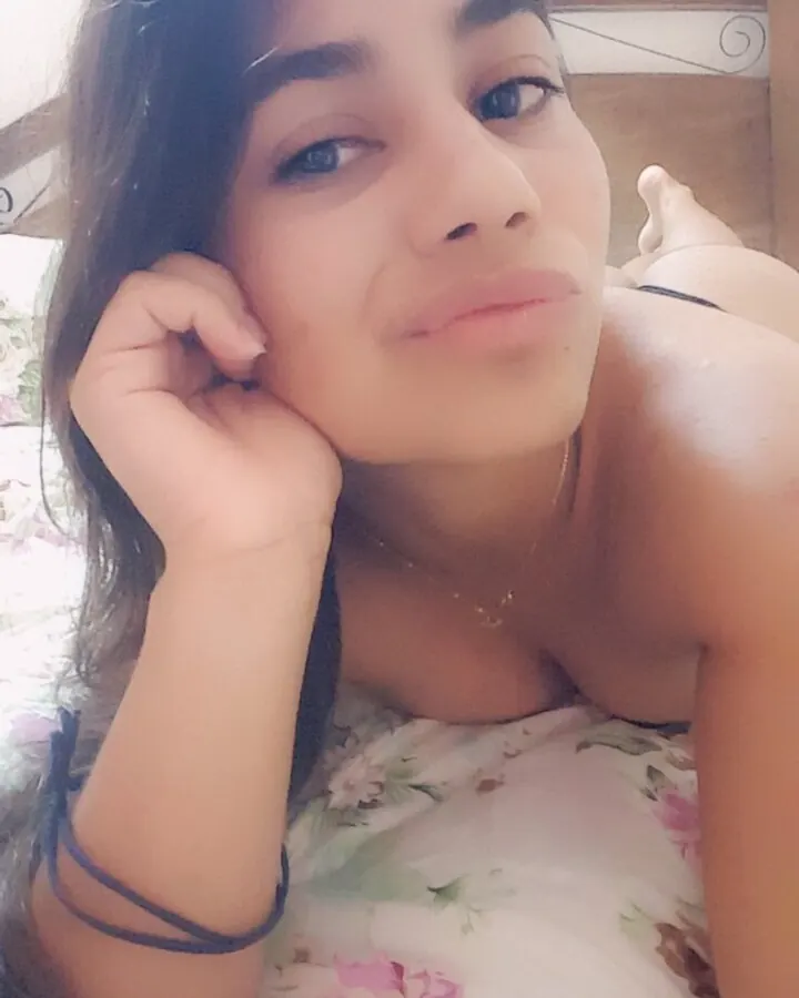 Jeune femme arabe aux yeux noirs, souriant légèrement, allongée sur un lit, portant un haut à motifs floraux blancs et roses.