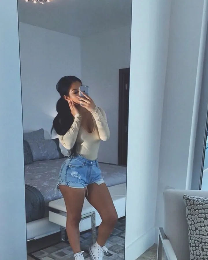 Jeune femme brune en body beige et short en jean, selfie dans une chambre