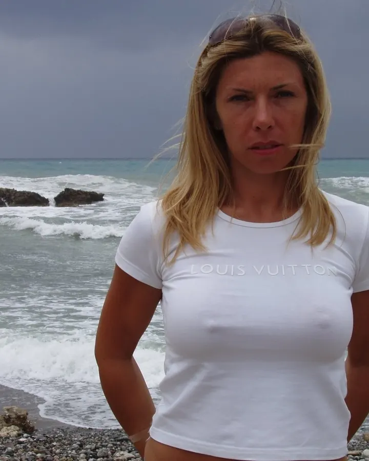 Femme en t-shirt blanc, debout sur une plage rocheuse, le regard fixe, sous un ciel gris