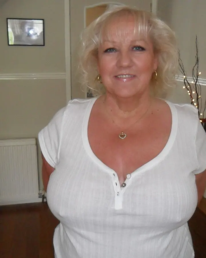 Femme aux cheveux blancs, souriante, en t-shirt blanc, debout dans une pièce lumineuse