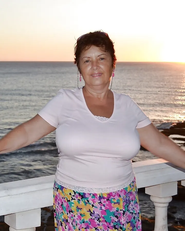 Femme souriante en tenue colorée, debout sur un pont face à la mer au coucher du soleil