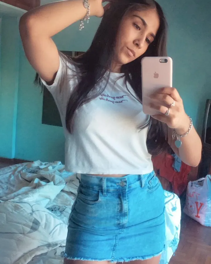 Jeune femme aux longs cheveux noirs, t-shirt blanc et jupe en jean, tenant un smartphone