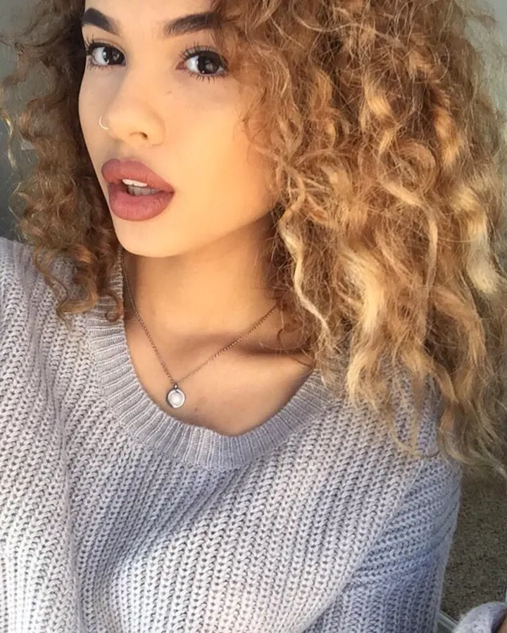 Jeune femme aux cheveux frisés blonds, pull gris, regard vers la caméra