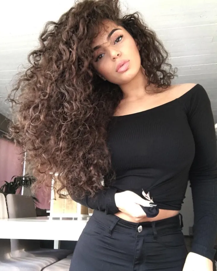 Femme aux cheveux châtains, portant un haut noir et un short en cuir, tenant un verre de vin, debout sur un sol en bois devant une porte en bois foncé.