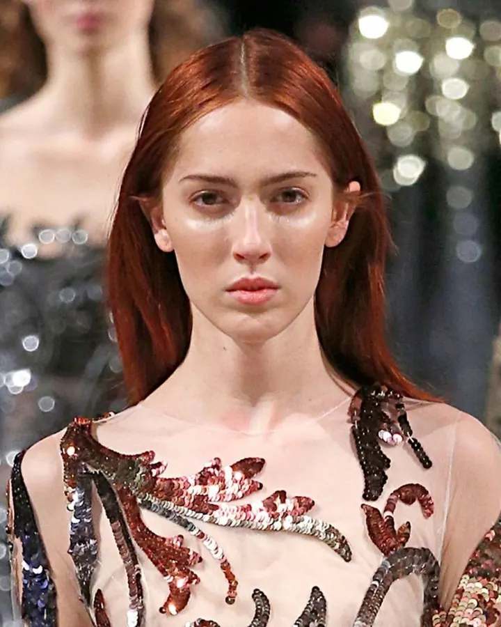 Mannequin aux cheveux roux portant une robe transparente ornée de sequins colorés sur un podium illuminé.