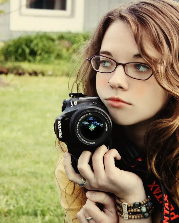 Jeune femme aux cheveux châtains et lunettes, tenant un appareil photo Pentax dans un jardin verdoyant.