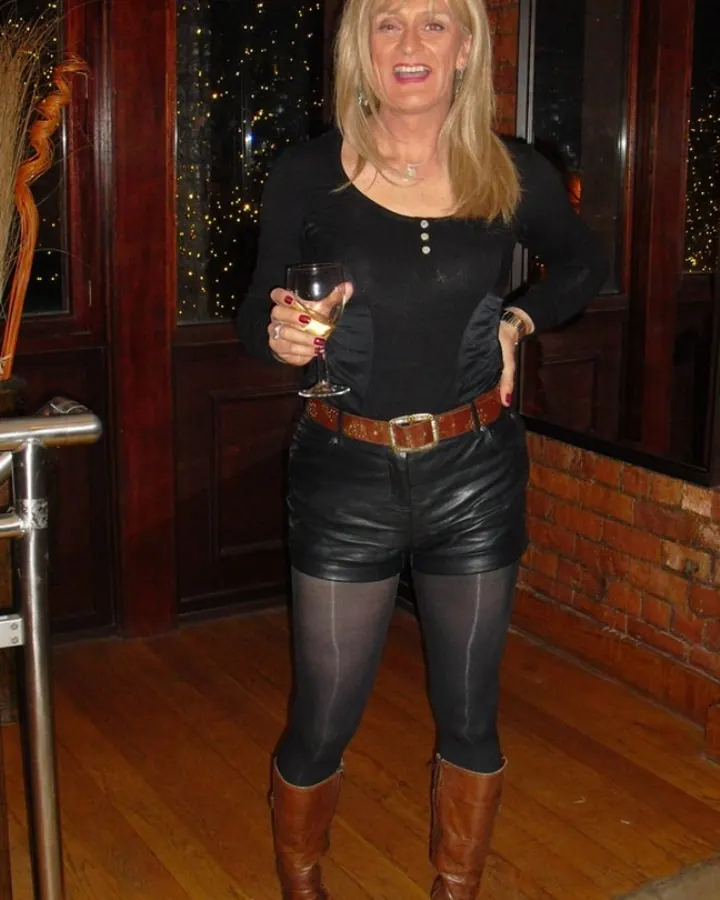 Femme aux cheveux blonds, portant un haut noir et un short en cuir, tenant un verre de vin, debout sur un sol en bois devant une porte en bois foncé.