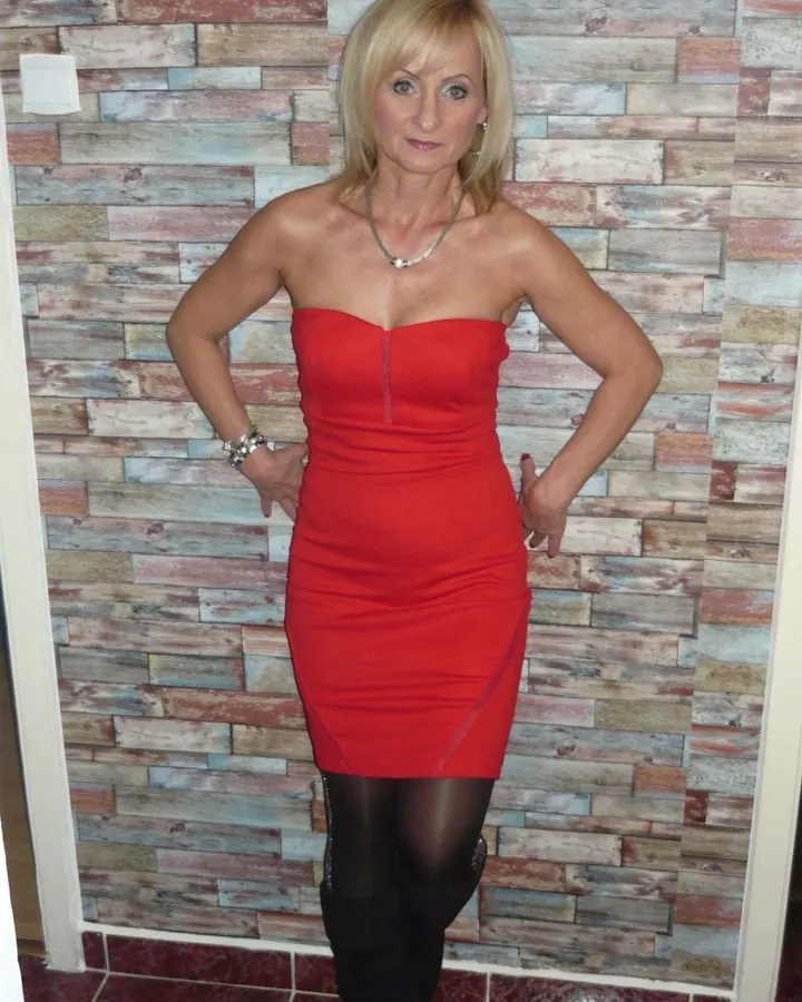 Femme aux cheveux blonds, souriante, en robe rouge, posant devant un mur en bois.