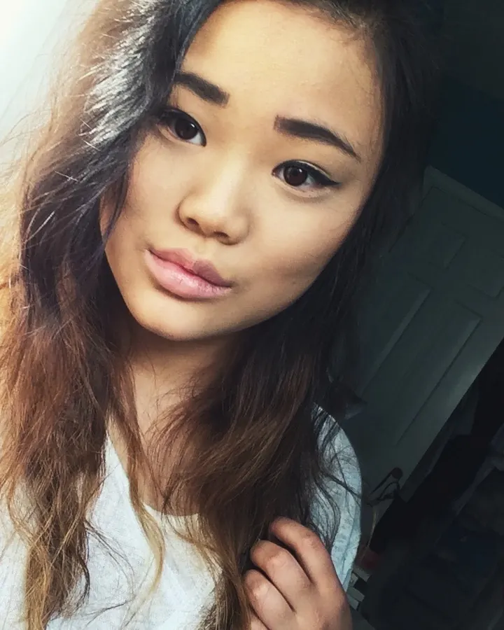 Jeune femme aux cheveux bruns, regardant la caméra, en haut blanc, dans une chambre.