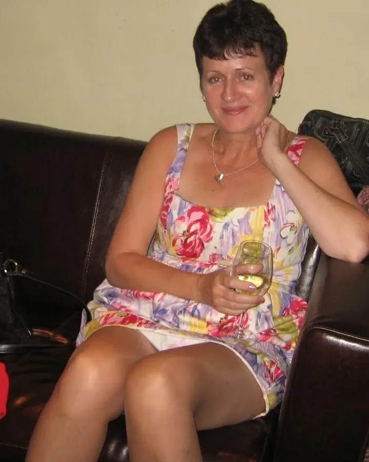 Femme aux cheveux bruns portant une robe blanche à fleurs, assise sur un canapé noir avec un verre de vin blanc dans la main.