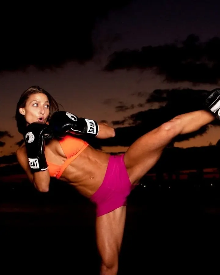 Femme en tenue de sport orange et rose, portant des gants de boxe, en train de frapper dans l'air contre un ciel crépusculaire.