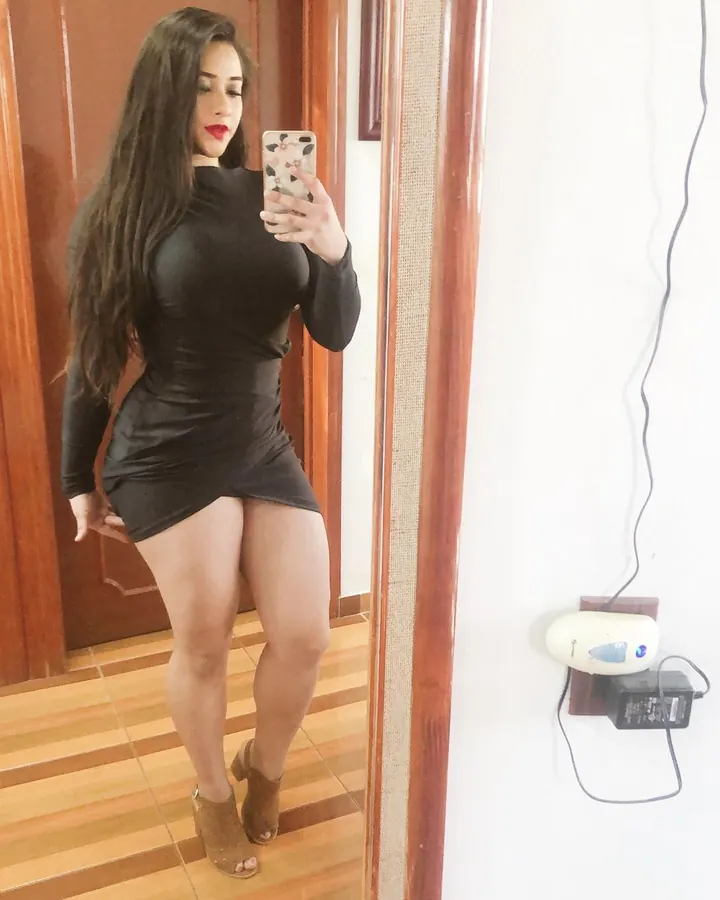 Femme aux cheveux longs noirs portant une robe noire moulante, prenant un selfie devant un miroir.