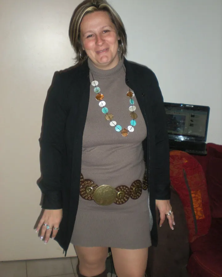 Femme aux cheveux bruns portant une robe noire, assise sur un plan de travail en cuisine avec des collants noirs et un blazer.