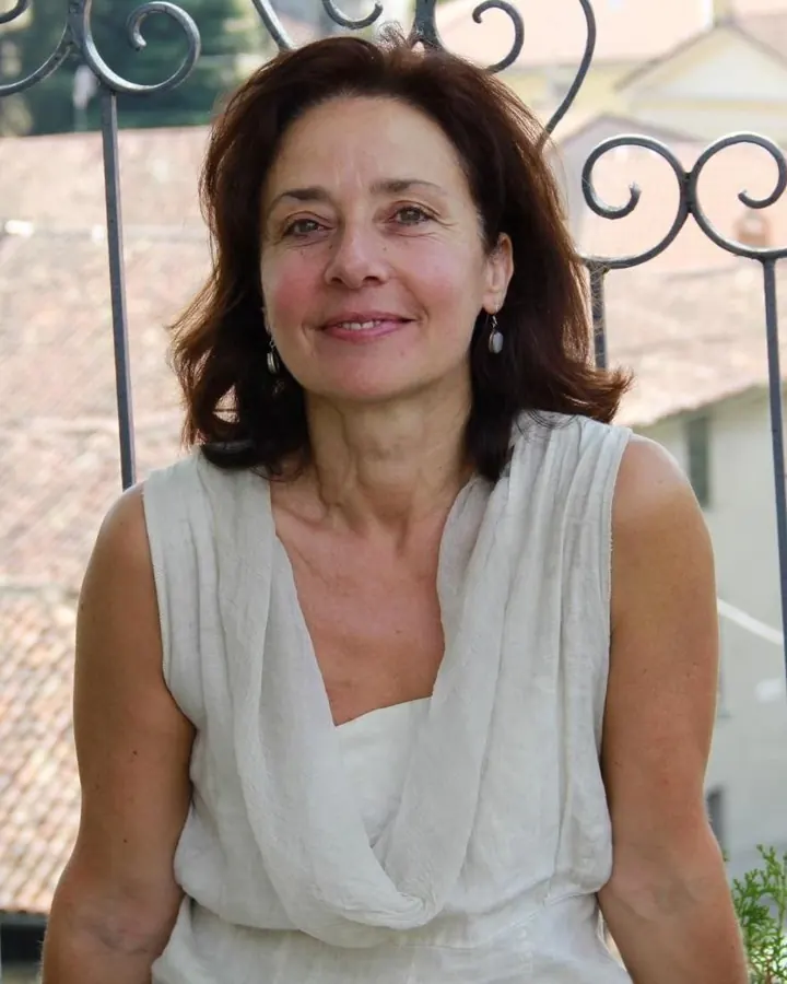 Femme aux cheveux bruns portant une robe blanche, souriante devant une grille métallique en extérieur.
