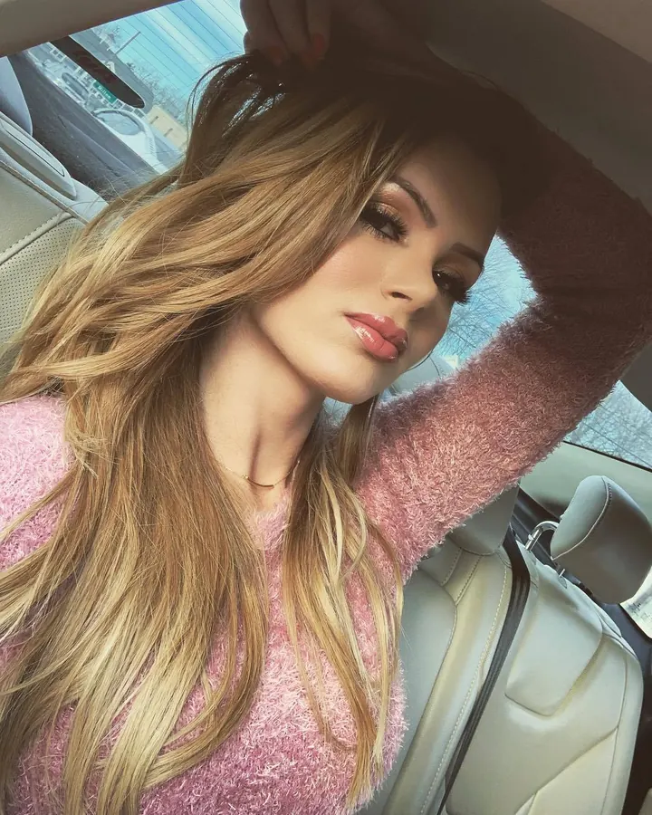 Femme blonde aux yeux bleus en pull rose dans une voiture, regardant vers l'appareil.