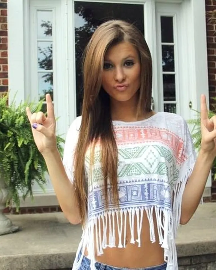 Femme brune en haut blanc, posant devant une maison en briques.